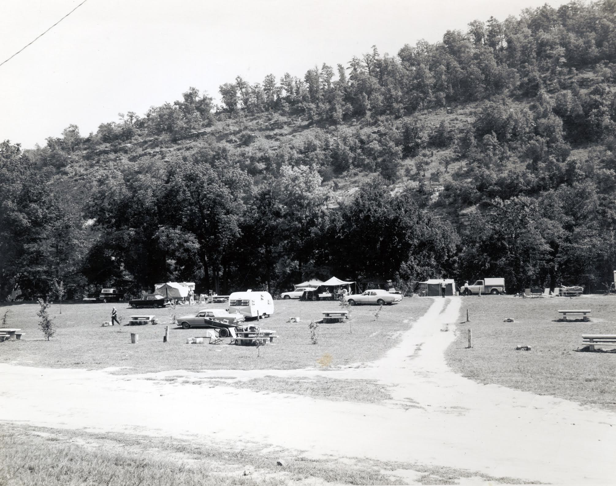 An open campground with RVs, tents, and cars from the 60s with trees in the background.
