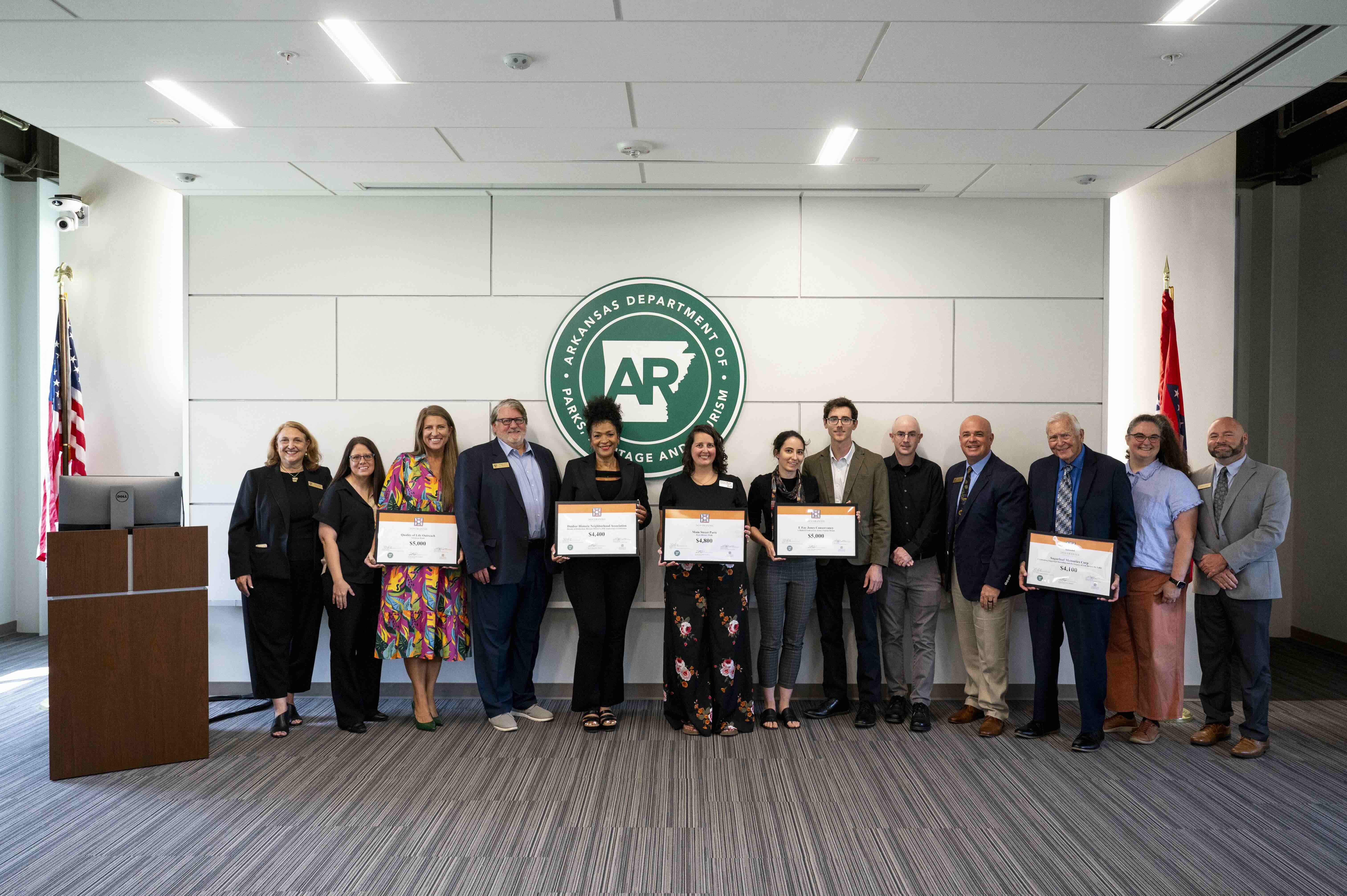 A group of grant recipients holding framed certificates posing in front of a large white wall.