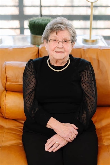 An older woman in a black dress and pearl necklace sitting on an orange couch.