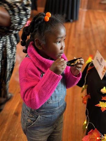 A young girl in jean overalls and a hot pink sweater eats a snack next to a table.